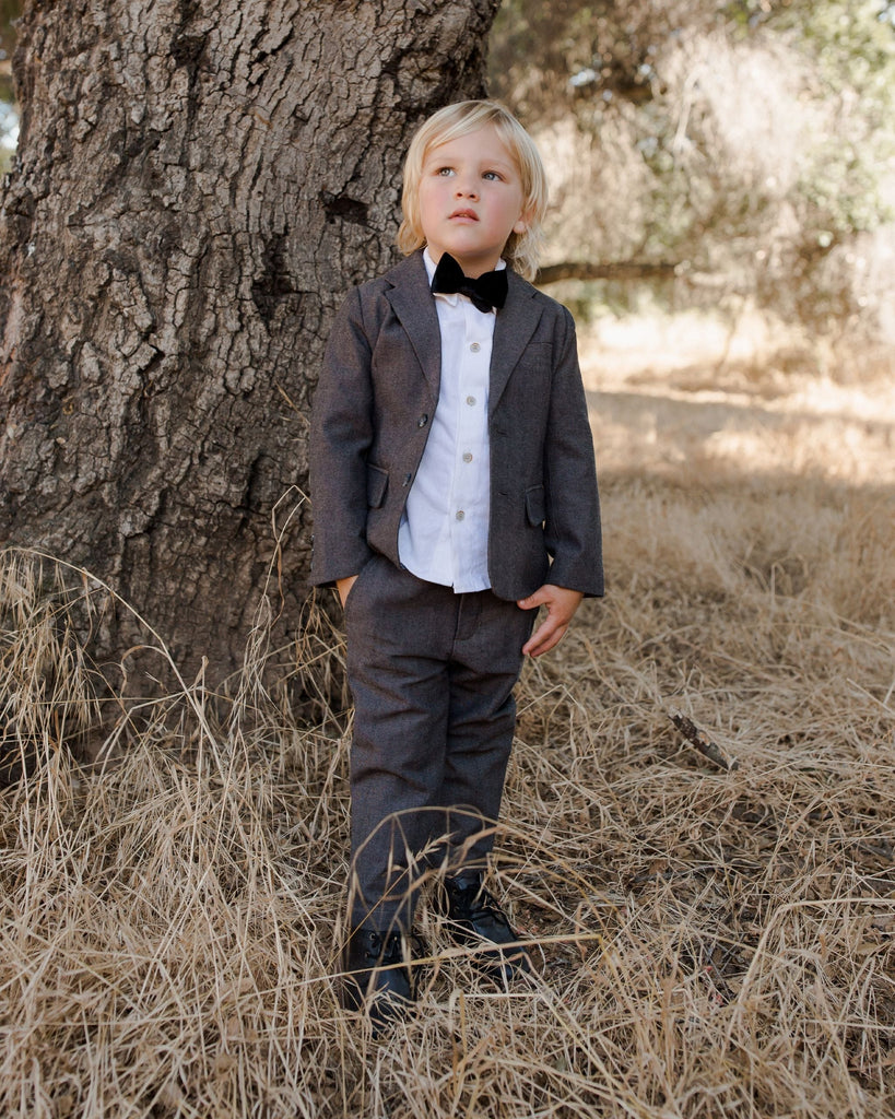 little boy outside wearing pants and jacket with bow time and tree behind 