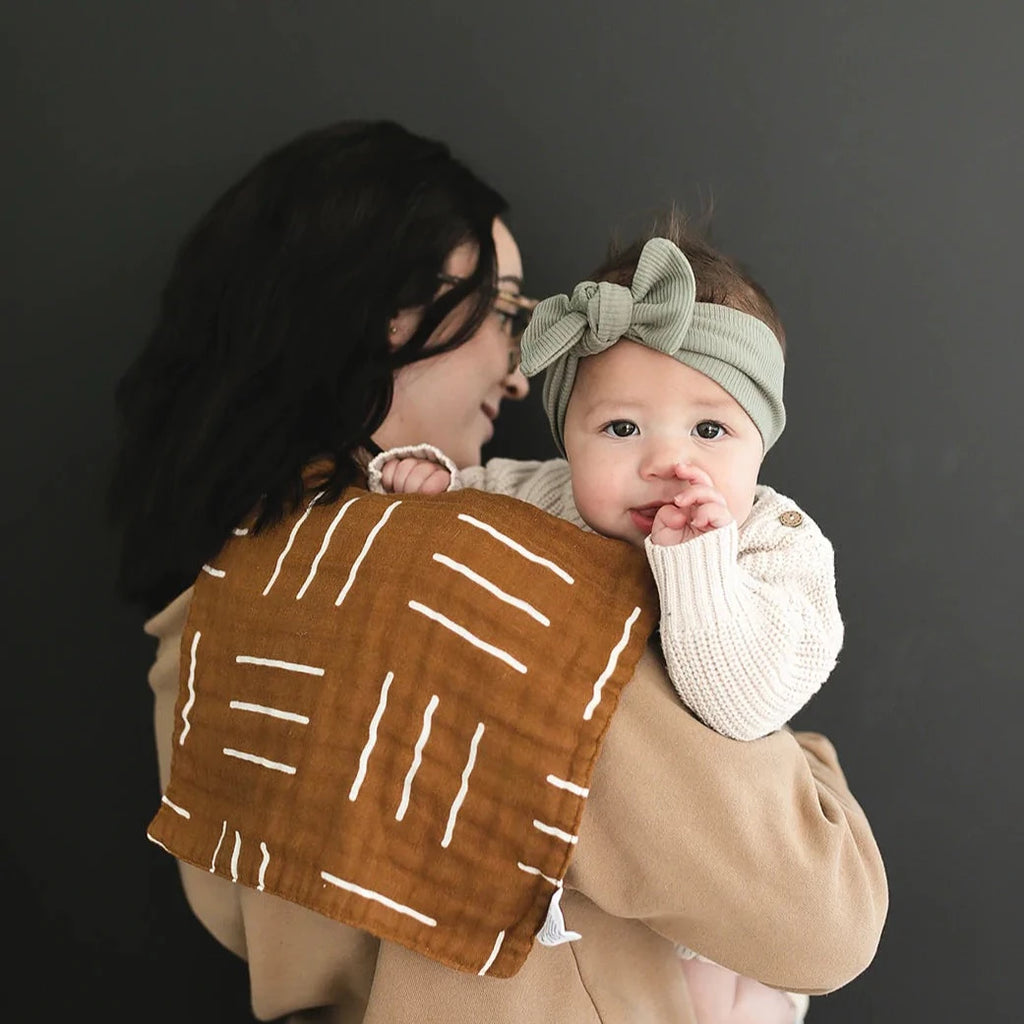 Dark background with mama holding baby, back facing camera, with a Mustard Mudcloth Burp Cloth by Mebie Baby draped over her shoulder. Burp Cloth is mustard with white lines all over.
