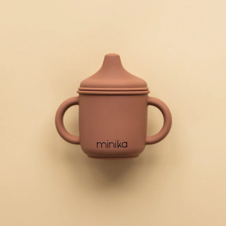 Beige background with a Silicone Sippy Cup in Cacao by Minika. Sippy cup is made of silicone, with 2 handles, a spouted lid, and the colour is dark brown and says “minika” in black.