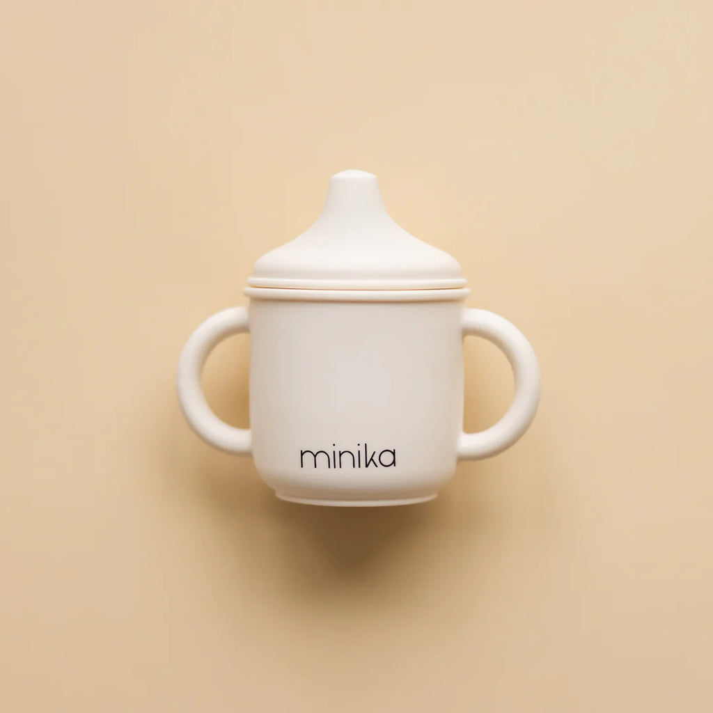 Beige background with a Silicone Sippy Cup in Shell by Minika. Sippy cup is made of silicone, with 2 handles, a spouted lid, and the colour is white and says “minika” in black.