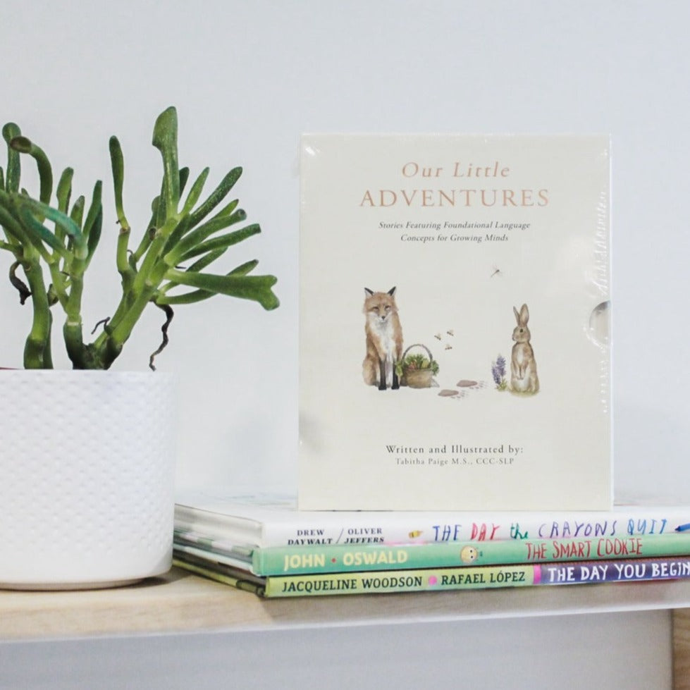 White wall with a natural wood shelf and a succulent, with a stack of books, and the book set Our Little Adventures: Stories Featuring Foundational Language Concepts for Growing Minds by Tabitha Paige. The box is cream with a watercolour fox, and bunny and a basket of veggies.