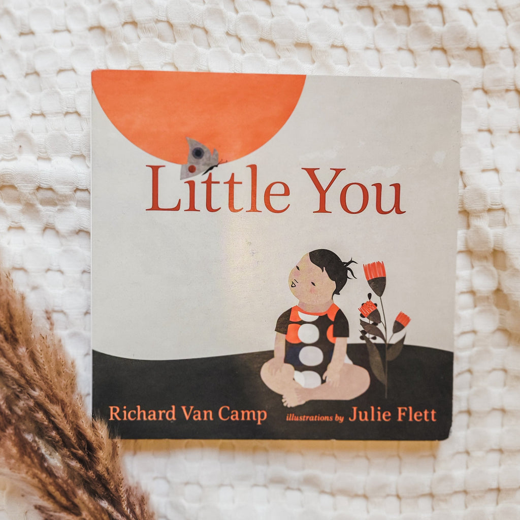 Cream waffle blanket with pampas grass on the bottom left corner, and the book Little You by Richard Van Camp. Cover is beige with a black ground,a nd a little person sitting cross legged beside some flowers.