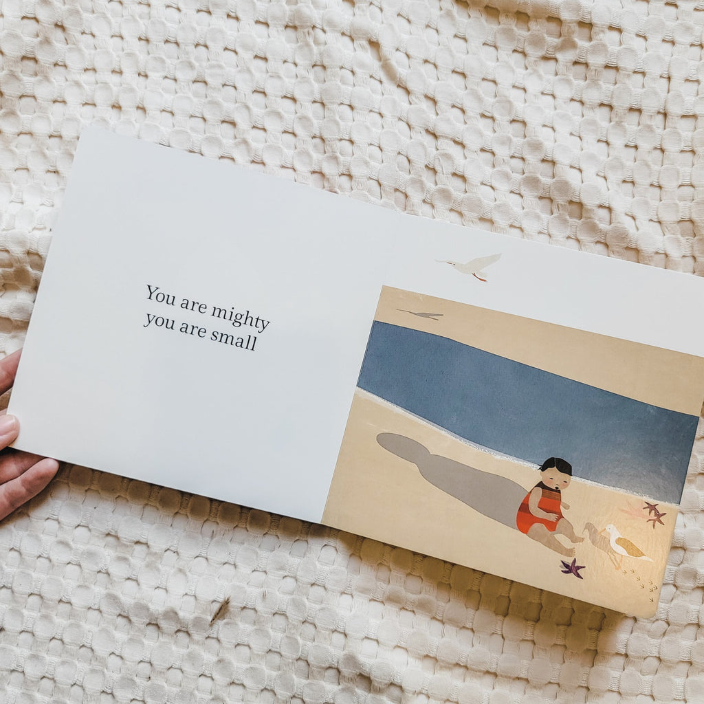 Cream waffle blanket with an open page from the book Little You by Richard Van Camp. Page is white on the left and says "You are mighty, you are small" and on the right it's a drawing of a little person sitting on the beach in the sand with a seagull.