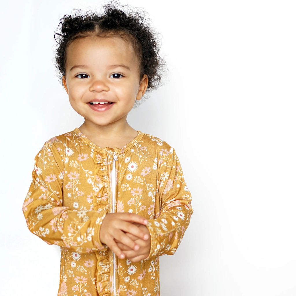 White background with toddler girl standing wearing the Bamboo Footie Sleeper in Mustard Floral by Emerson and Friends.