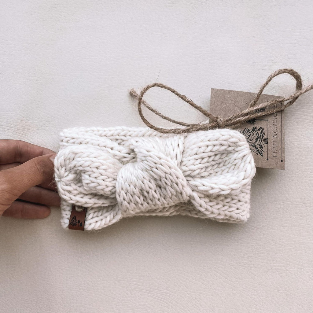 White background with a hand out, holding the Handknit Knot Bow Headband by Petit Nordique in Natural. Natural is a creamy white yarn, and its got a knot detail on the front and a small leather tag.