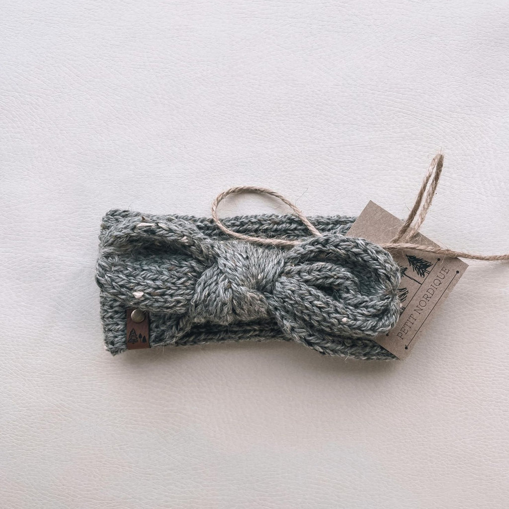 White background with a Handknit Knot Bow Headband by Petit Nordique in Grey Tweed. Headband has a knot bow in the front, with a small leather tag, in a grey tweed yarn.