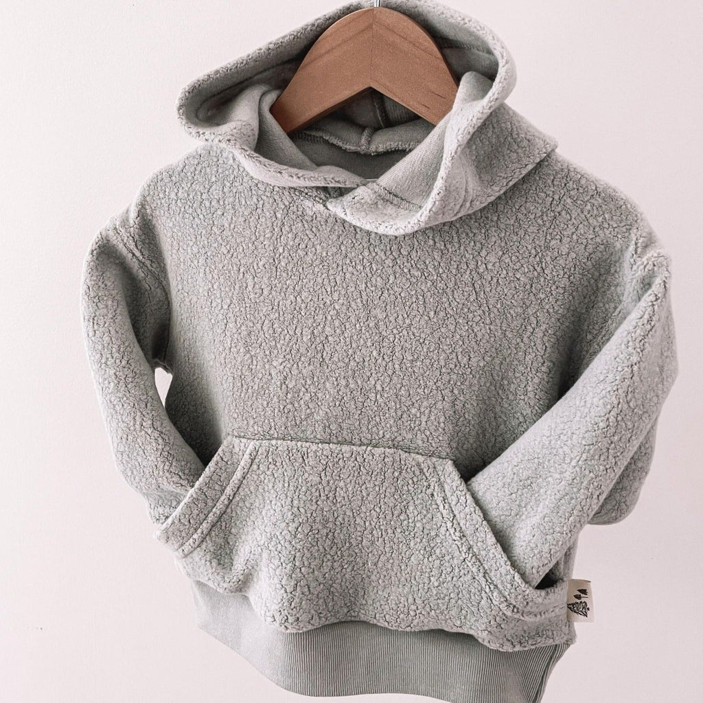 Grey Winter Sky Hoodie by Petit Nordique hanging on a wooden hanger in front of a white wall. 