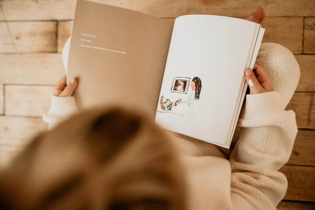 Overhead view of a little girl sitting on a wood floor, with a page open from the book 'Worth The Wait" | Written & Illustrated by Andrée Linnell. The page is white and has a drawing of a woman laying down getting an ultrasound.
