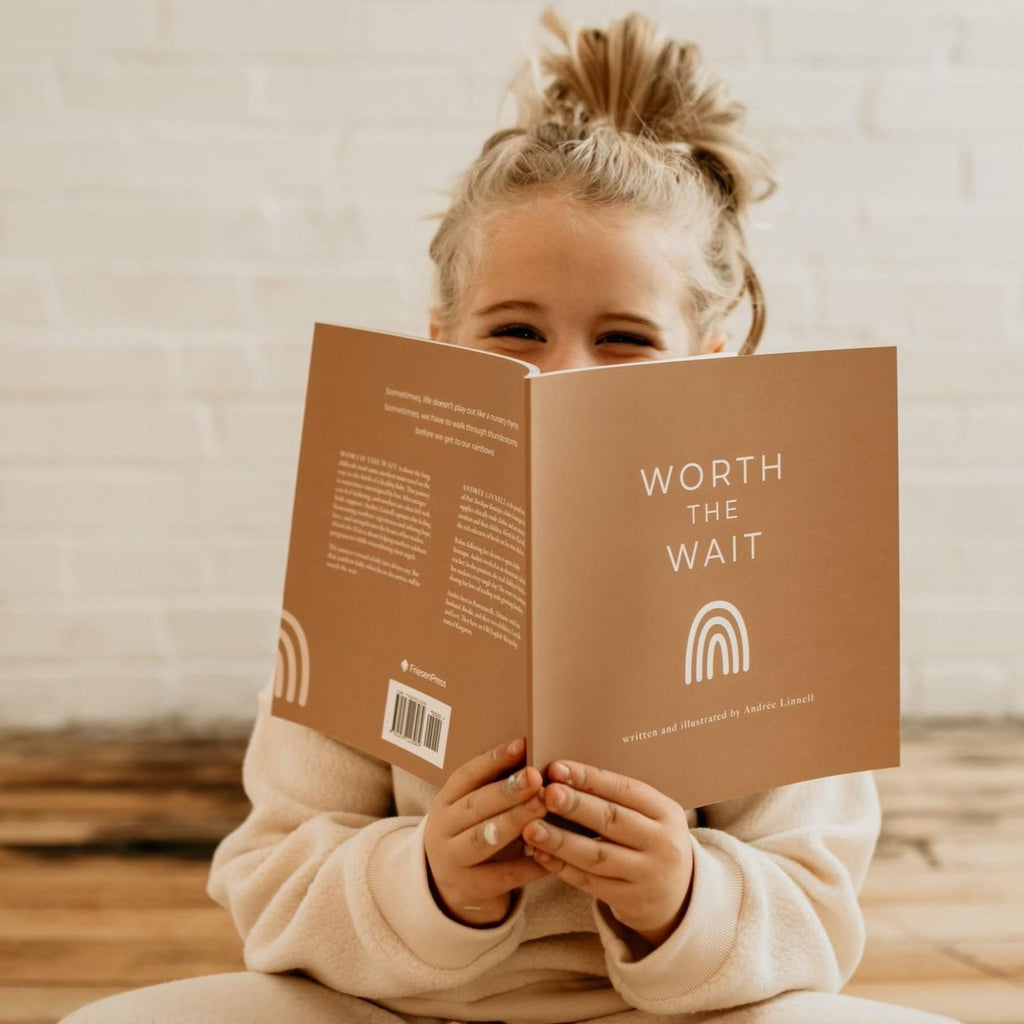 White brick wall with natural wood floors and a little girl sitting, holding the 'Worth The Wait' Book | Written & Illustrated by Andrée Linnell in front of her face. Cover is brown and says "Worth the wait" in white, with a white rainbow.