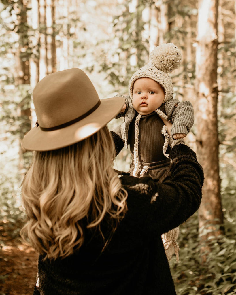 Back view of a woman standing, holding her baby in the air with trees behind, wearing The Nordique Women's Shacket 2.0 in Black by Petit Nordique.