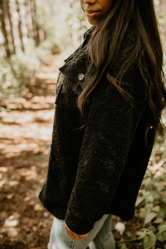 Woman standing with trees behind her, to the side, showing a side view of The Nordique Women's Shacket 2.0 in Black by Petit Nordique.