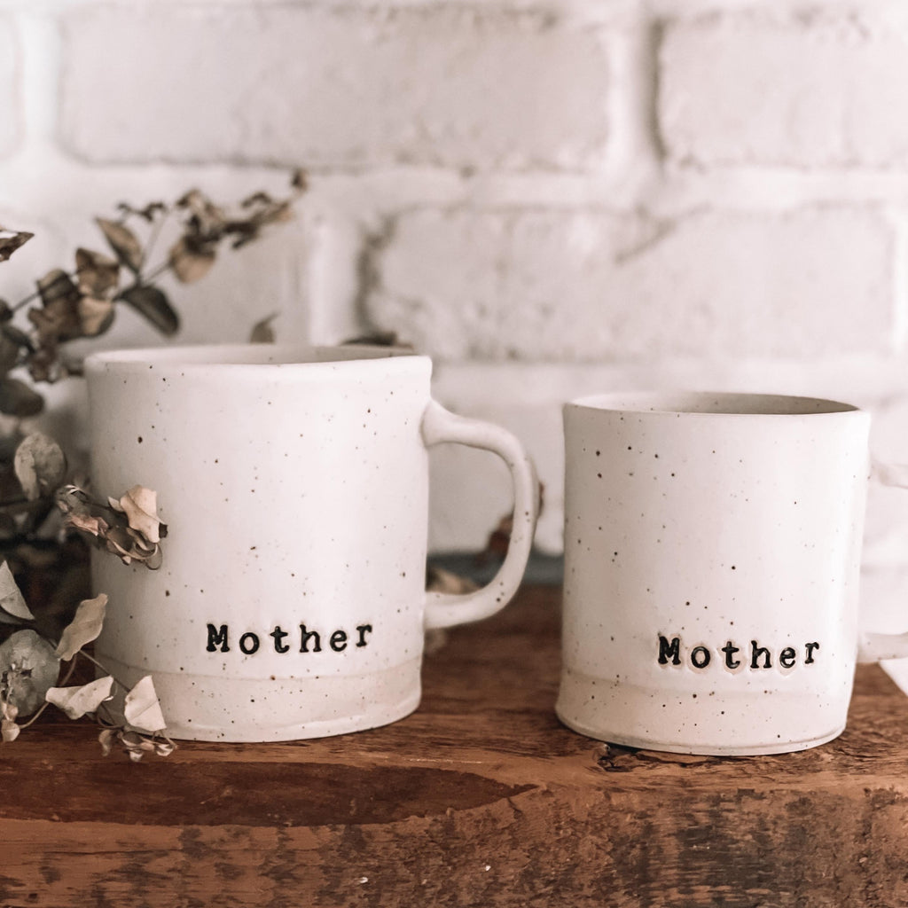 White brick wall with a wooden floating shelf, with the Mother Handmade Mug by Beech Hill Studio. Showing both mugs, 16oz and the 12oz.