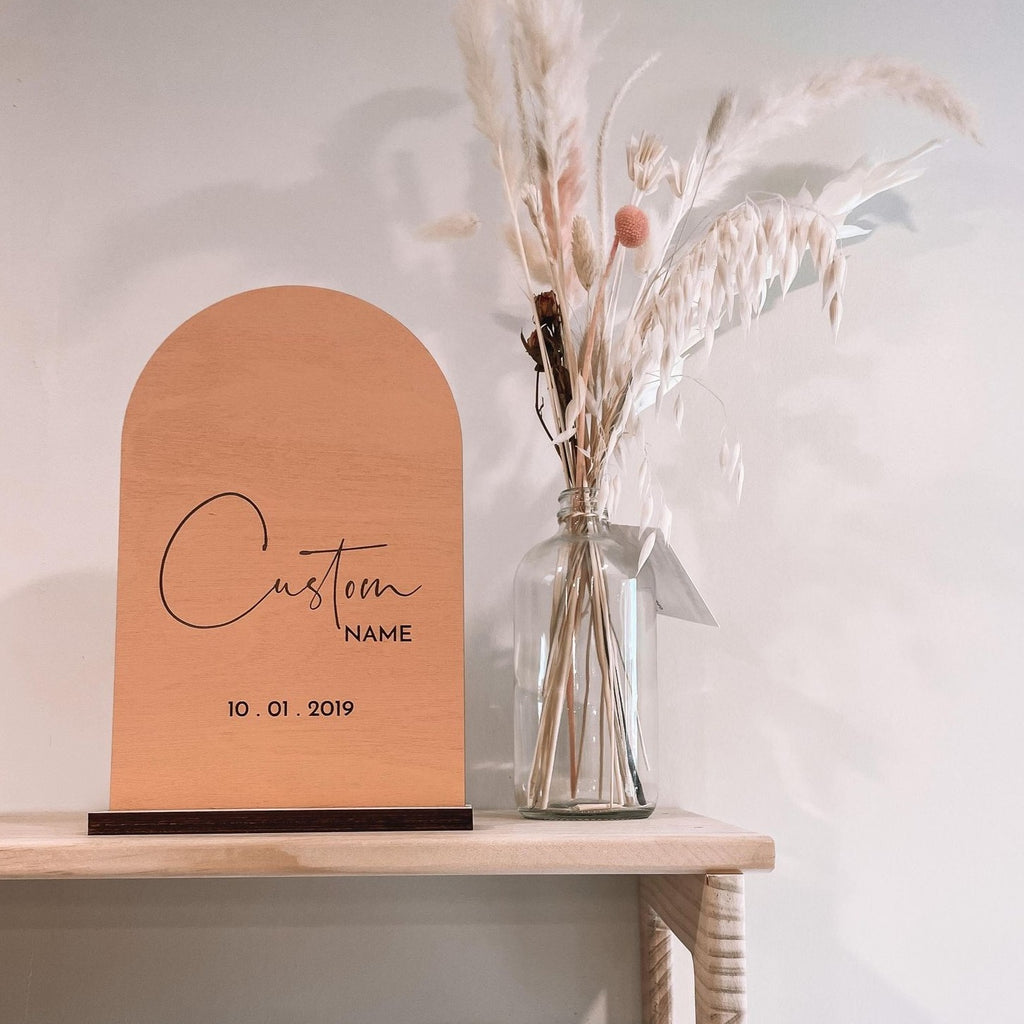 White wall with a natural wood shelf, and the Personalized Name Arch in Parfait by Petit Nordique beside a jar of pampas grass. Name arch is an arch, in a coral/peach colour with custom writing on a wooden base.