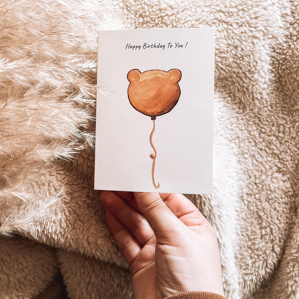 Bear Balloon | Birthday card by PARKES & Bash, being held up by a hand. In background, there is pampas grass and a beige sherpa blanket by Petit Nordique. 