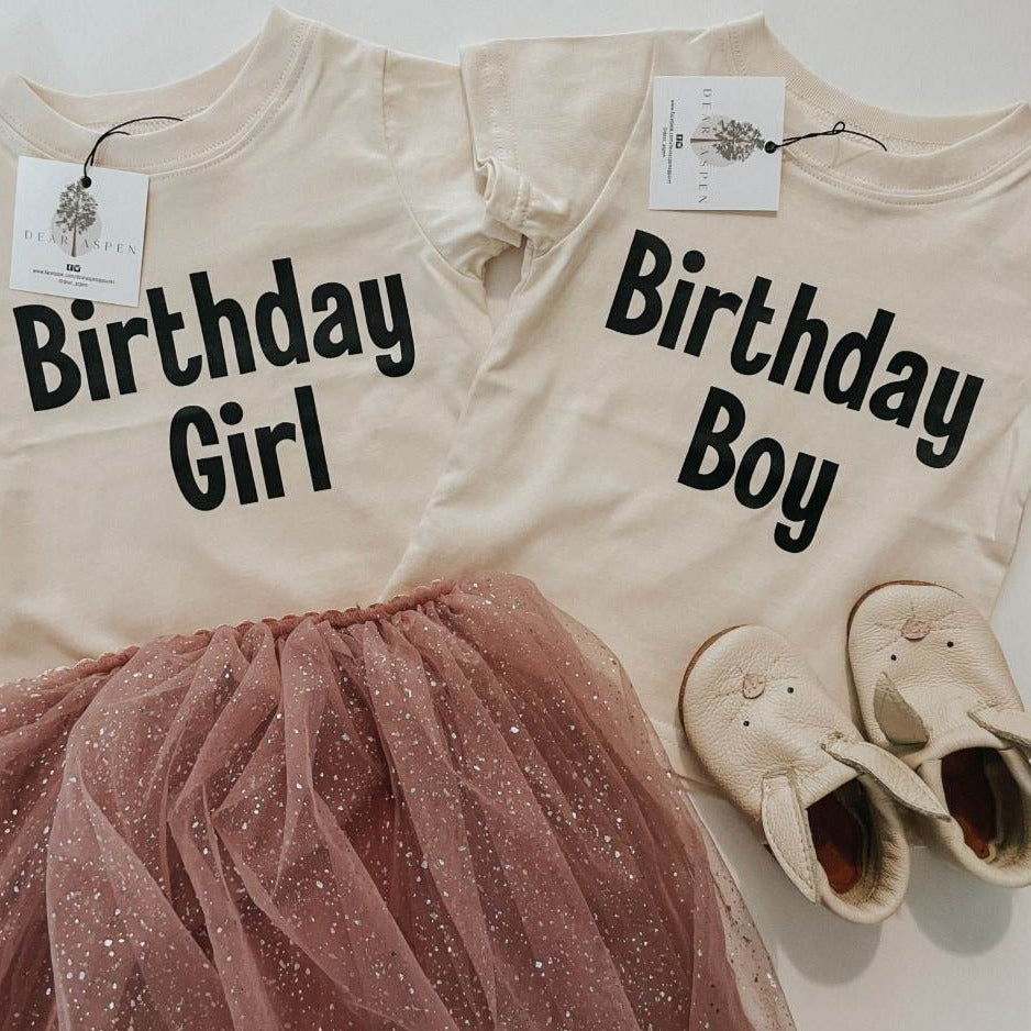 Birthday Boy and Birthday Girl Tees by Dear Aspen. Birthday Girl Tee paired with pink tutu and Birthday Boy Tee paired with bunny moccs, laid on a flat white surface. 