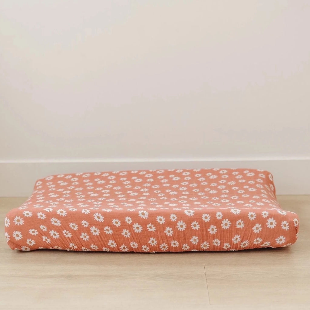 White wall with light wood floor, and a changing pad with the Arizona Daisy Changing Pad Cover by Mebie Baby on it. Changing pad cover is orange with daisies all over, and fits snug.