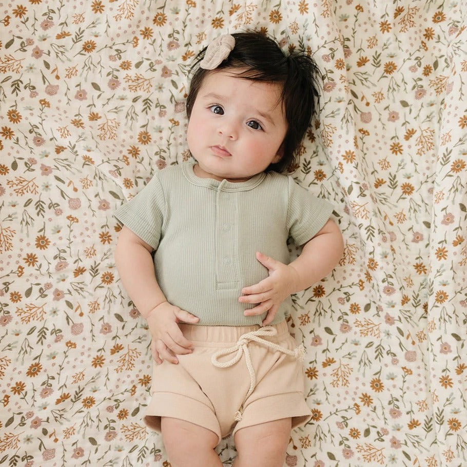 Overhead view of baby girl laying on her back, wearing a Sage Organic Cotton Ribbed Snap Bodysuit by Mebie Baby. Bodysuit is sage, ribbed, and has 3 buttons down the front.