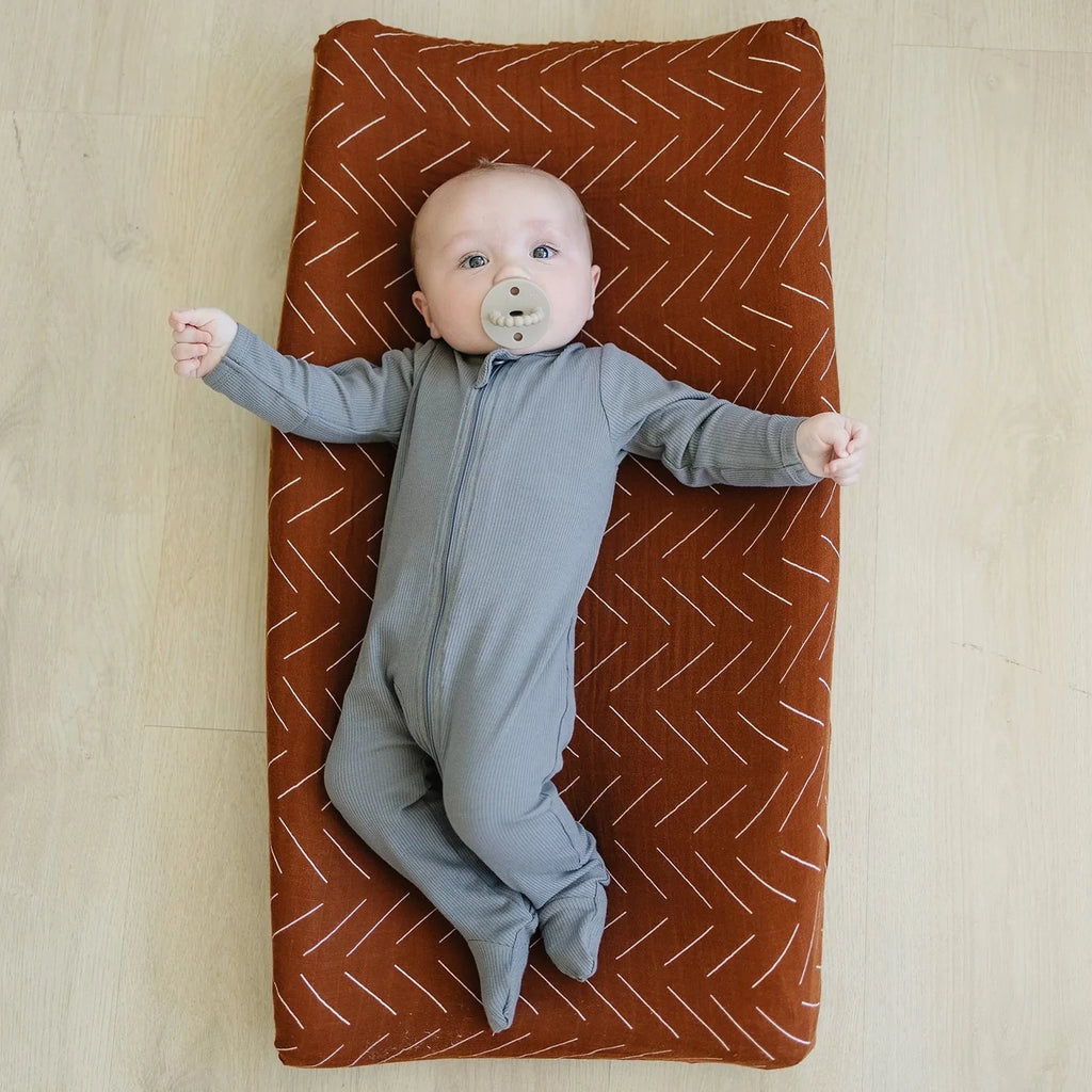 Light wood floors with a baby laying on a change pad with a Rust Mudcloth Changing Pad Cover by Mebie Baby on it. Cover is rust with white lines all over.