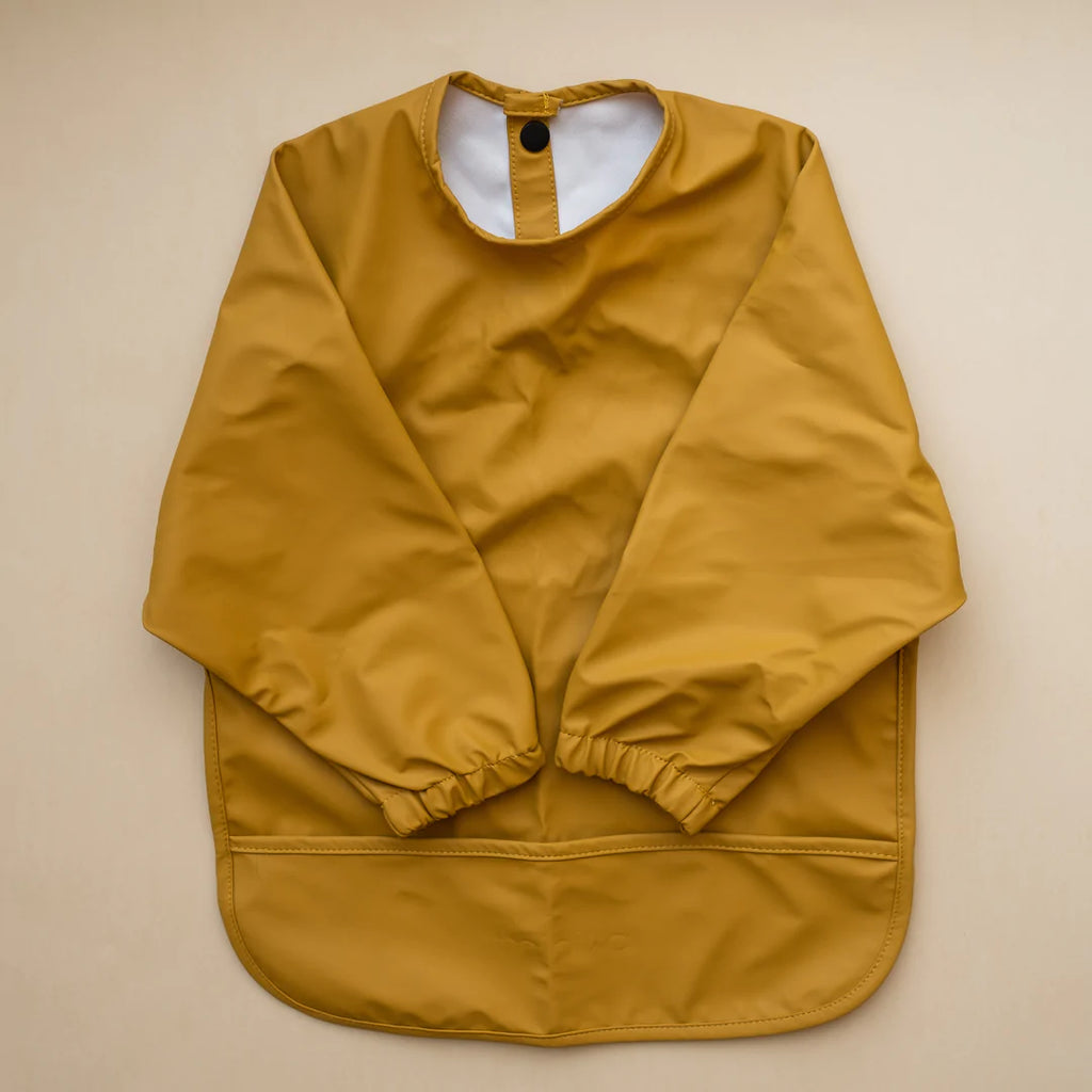 Beige background with a Long Sleeve Bib in Ochre by Minika. Bib is ochre with long sleeves, snaps on the back, and a pouch along the bottom.