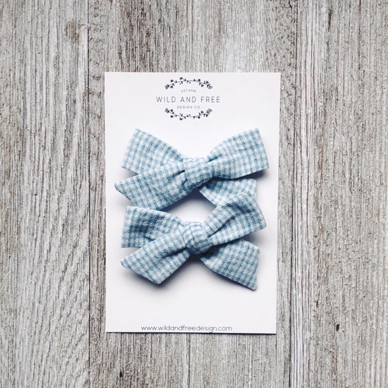 Overhead view of the Blue and White Check School Girl Pigtail Bow Set by Wild and Free Design Co., on a grey washed table. Set has 2 bows, in a white and blue checkered pattern.