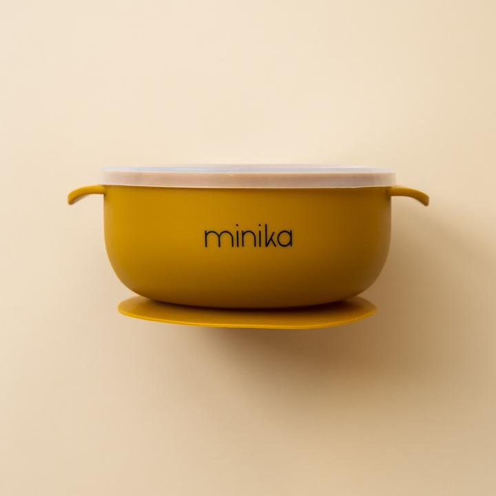 Beige background with a Silicone Bowl with Lid in Ochre by Minika. Bowl is mustard/ochre, has a silicone base to stick to your table, and a clear lid.