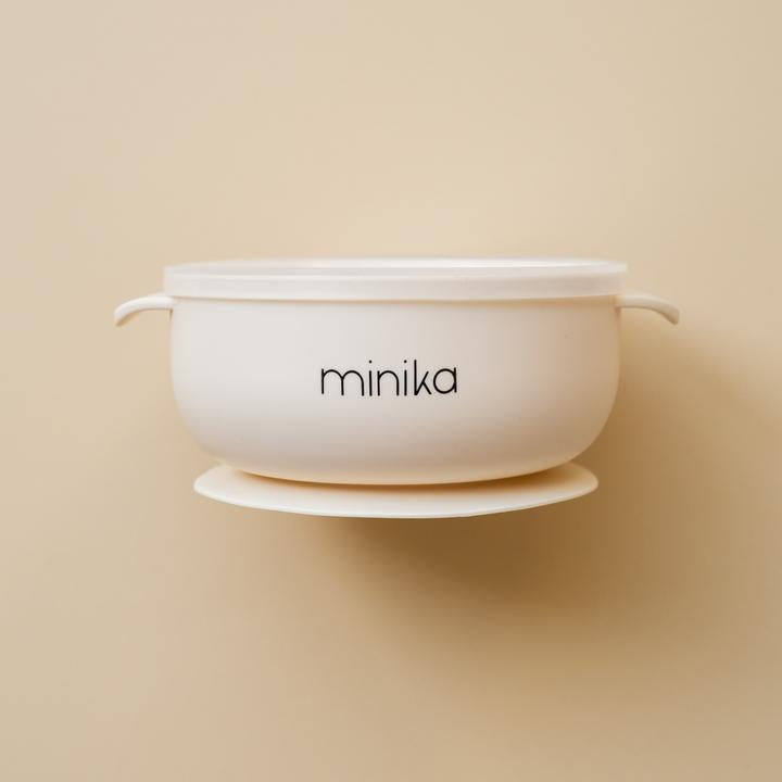 Beige background with a Silicone Bowl with Lid in Shell by Minika. Bowl is white, has a silicone base to stick to your table, and a clear lid.