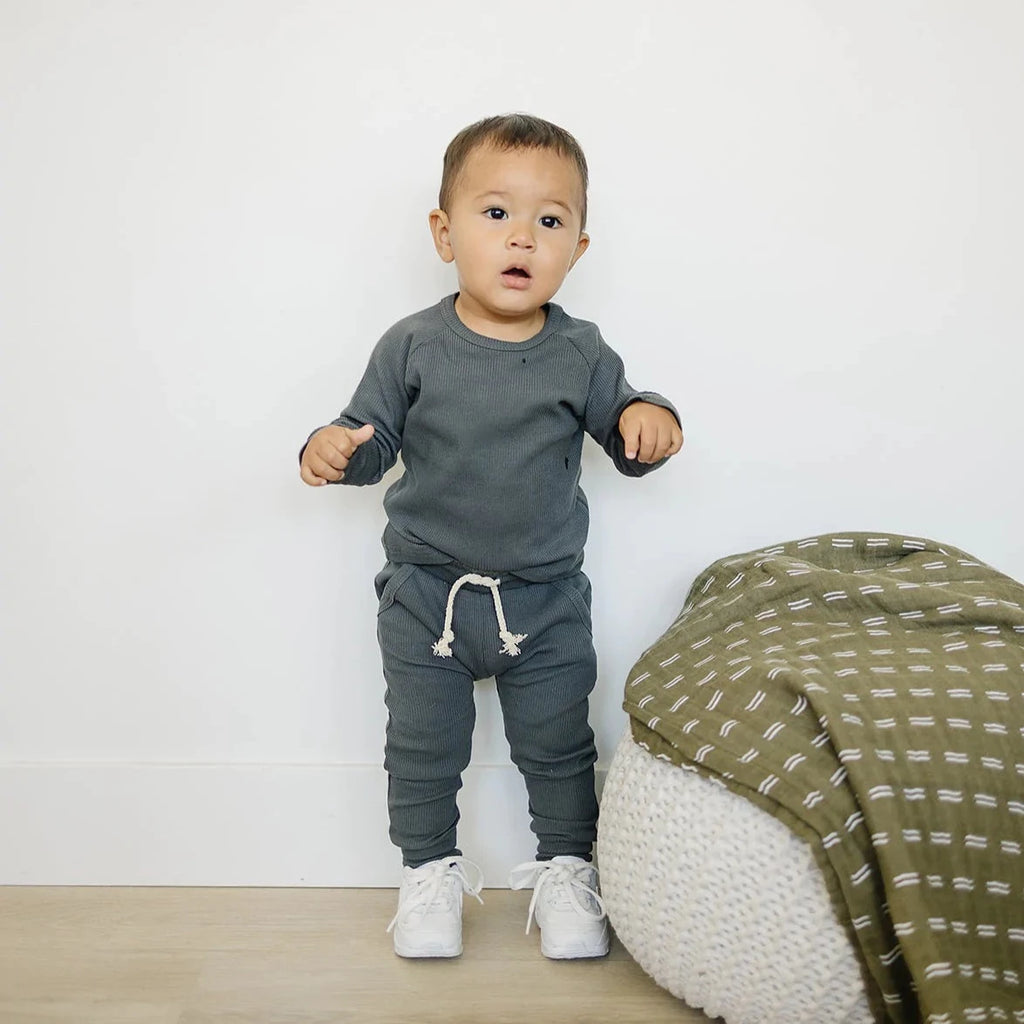 White wall with a white pouf, and an Olive Pines Quilt on it, and a toddler boy standing wearing the Charcoal Organic Cotton Ribbed Pocket Set by Mebie Baby.