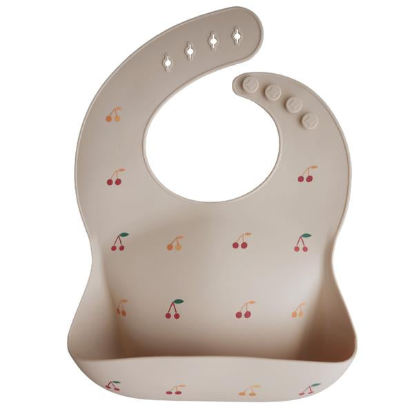 White background with a Silicone Bib in Cherries by Mushie. Bib is a beige colour with red & orange cherries, with a deep pocket on the front, and a rounded neck fastener at the back.