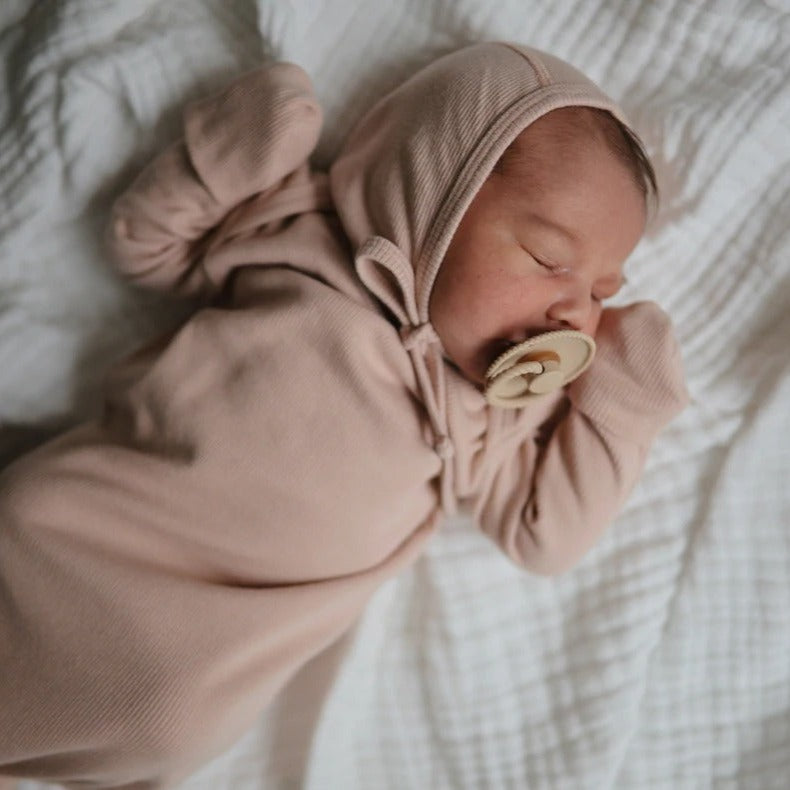 Overhead view of a baby sleeping on a white muslin blanket, wearing a Ribbed Baby Bonnet by Mushie.