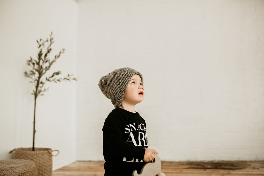 White brick wall with a little boy standing, looking up and wearing a Handknit Beanie by Petit Nordique in Grey Tweed.