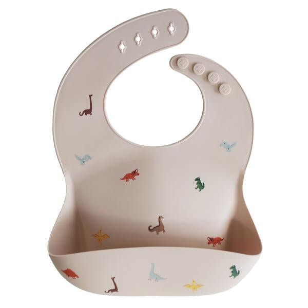 White background with a Silicone Bib in Dinosaurs by Mushie. Bib is a beige colour with colourful dinosaurs, with a deep pocket on the front, and a rounded neck fastener at the back.