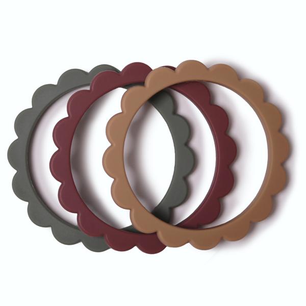 White background with Flower Teething Bracelet 3-Pack in Dried Thyme/Berry/Natural by Mushie. These are round bracelets, that look like flower petals - comes with a dark green, reddish, and brown bracelet.
