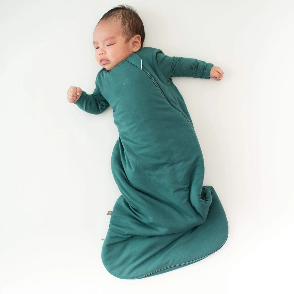 White background with overhead view of baby laying down, wearing a Sleep Bag 2.5 Tog in Emerald by Kyte Baby. Sleep bag is an emerald green, with a side zipper going all around the bottom.