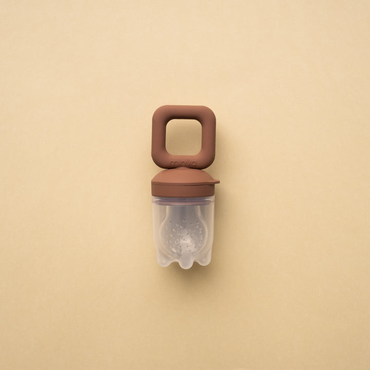 Beige background with a Silicone Feeder Teether in Cacao by Minika. Feeder teether has a dark brown silicone handle, and the feeder part is clear silicone with small holes.