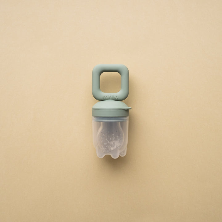 Beige background with a Silicone Feeder Teether in Sage by Minika. Feeder teether has a sage silicone handle, and the feeder part is clear silicone with small holes.