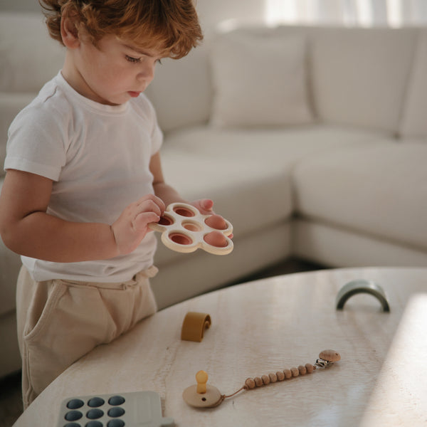 Living room, with a white sectional, and a close up view of a boy standing at a natural wood table playing with the Flower Press Toy by Mushie. 