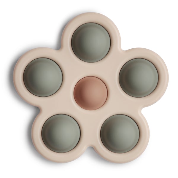 White background with Flower Press Toy in Dried Thyme/Natural/Shifting Sand by Mushie. This is a beige colour silicone shaped like a 5 point flower, with 5 green "pop its" around the outside, and 1 brown in the center.
