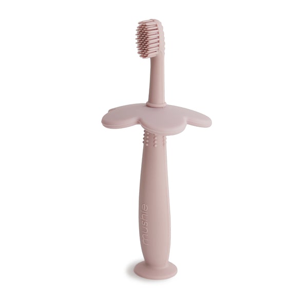 White background with Flower Training Toothbrush in Blush by Mushie. Toothbrush is made out of silicone with a flower style safety guard, in a blush colour.