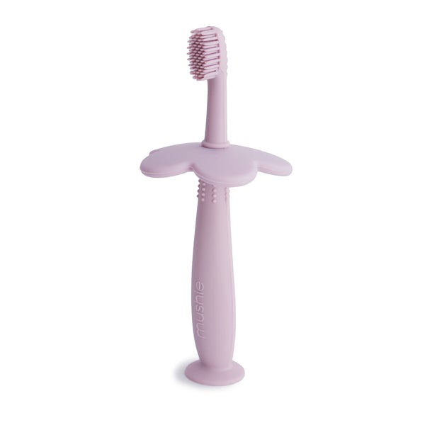 White background with Flower Training Toothbrush in Soft Lilac by Mushie. Toothbrush is made out of silicone with a flower style safety guard, in a lilac colour.