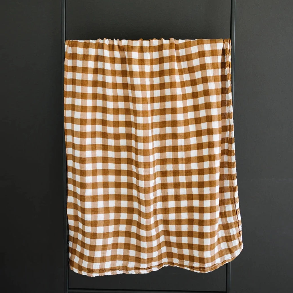 Dark background with a metal blanket ladder, and a Gingham Muslin Swaddle by Mebie Baby hanging from it. This swaddle is brown and white gingham pattern.