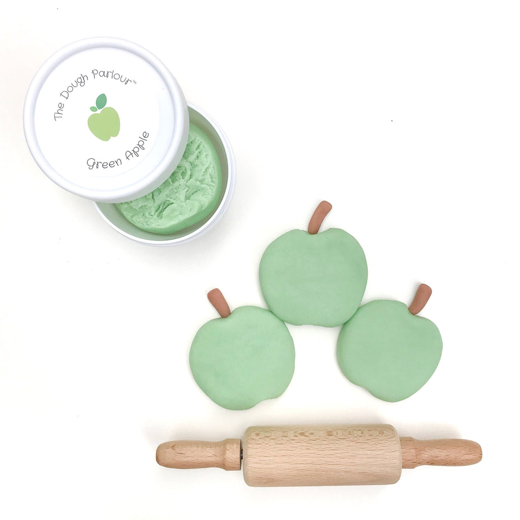 White background with open container of Green Apple Play Dough by Dough Parlour in the top left, with 3 little green apples made out of dough and a rolling pin.