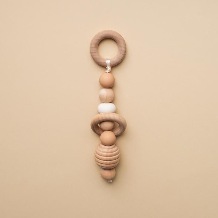 Beige background with a Hand Rattle in Natural by Minika. Hand rattle has a wooden circle with beige silicone, white silicone and wood beads going down, and a honeycomb bead.