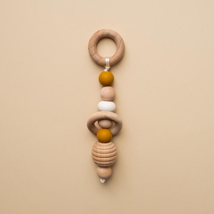 Beige background with a Hand Rattle in Ochre by Minika. Hand rattle has a wooden circle with mustard silicone, white silicone and wood beads going down, and a honeycomb bead.