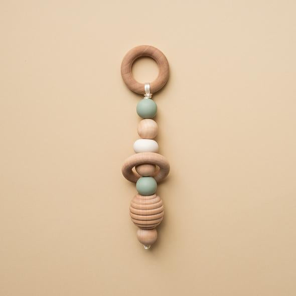 Beige background with a Hand Rattle in Sage by Minika. Hand rattle has a wooden circle with sage silicone, white silicone and wood beads going down, and a honeycomb bead.