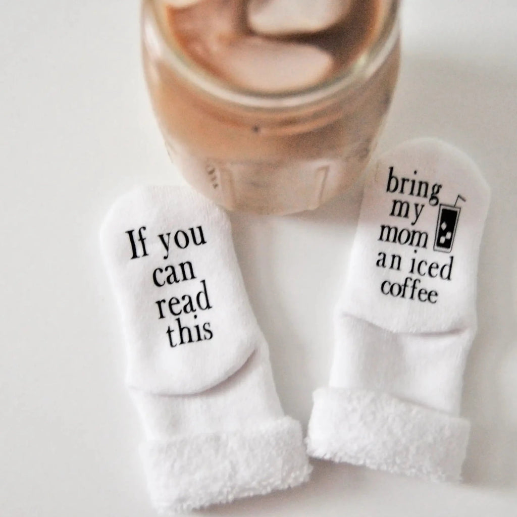 White background with an iced coffee, and a pair of Buy My Mom An Iced Coffee Baby Socks by Dorothy's Reason. Socks are white, one says "If you can read this", other one says "Bring my mom an iced coffee".