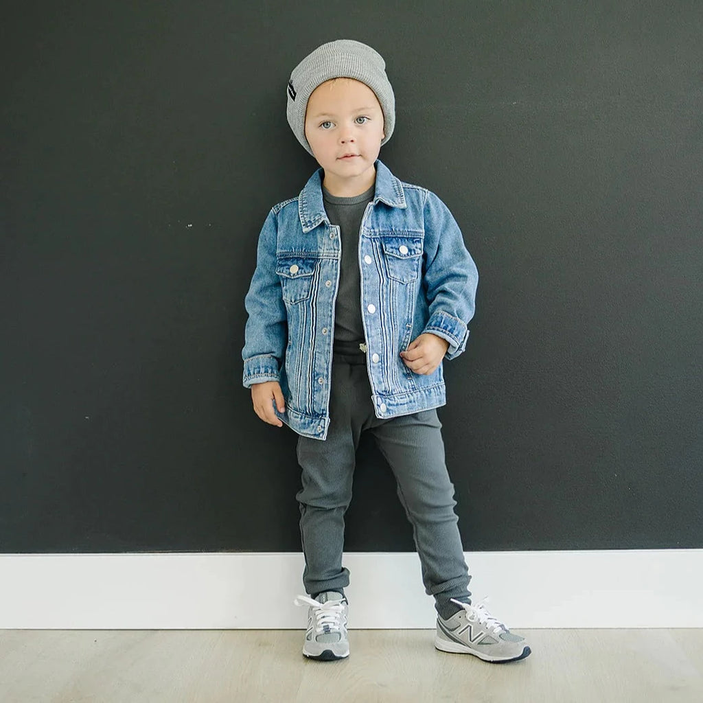 Dark background with a toddler boy standing, wearing a two piece mebie set with a beanie, and the Mebie Baby Jean Jacket by Mebie Baby over top.