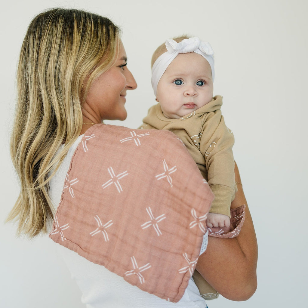 Just Peachy Burp cloth By Mebie Baby draped over mothers shoulder, while she's holding her baby. Mother is standing in front of a white wall. 