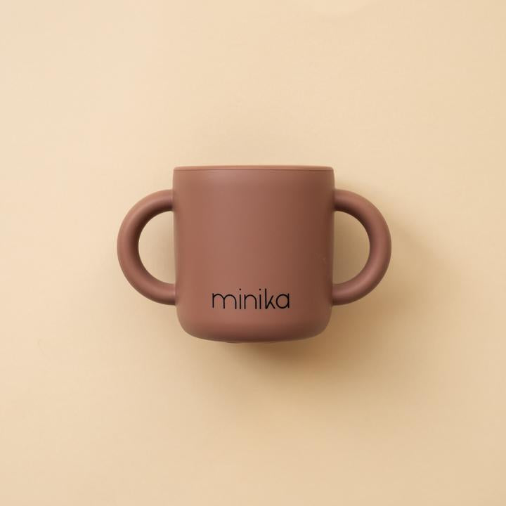 Beige background with a Learning Cup with Handles in Cacao by Minika. Cup is dark brown silicone with 2 handles, and says “minika” in black
