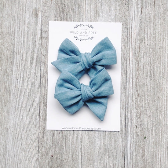 Overhead view of the Light Washed Denim Pigtail Bow Set by Wild and Free Design Co. on a white washed wood table, in its packaging. Bows are a large pinwheel style in a medium blue color.