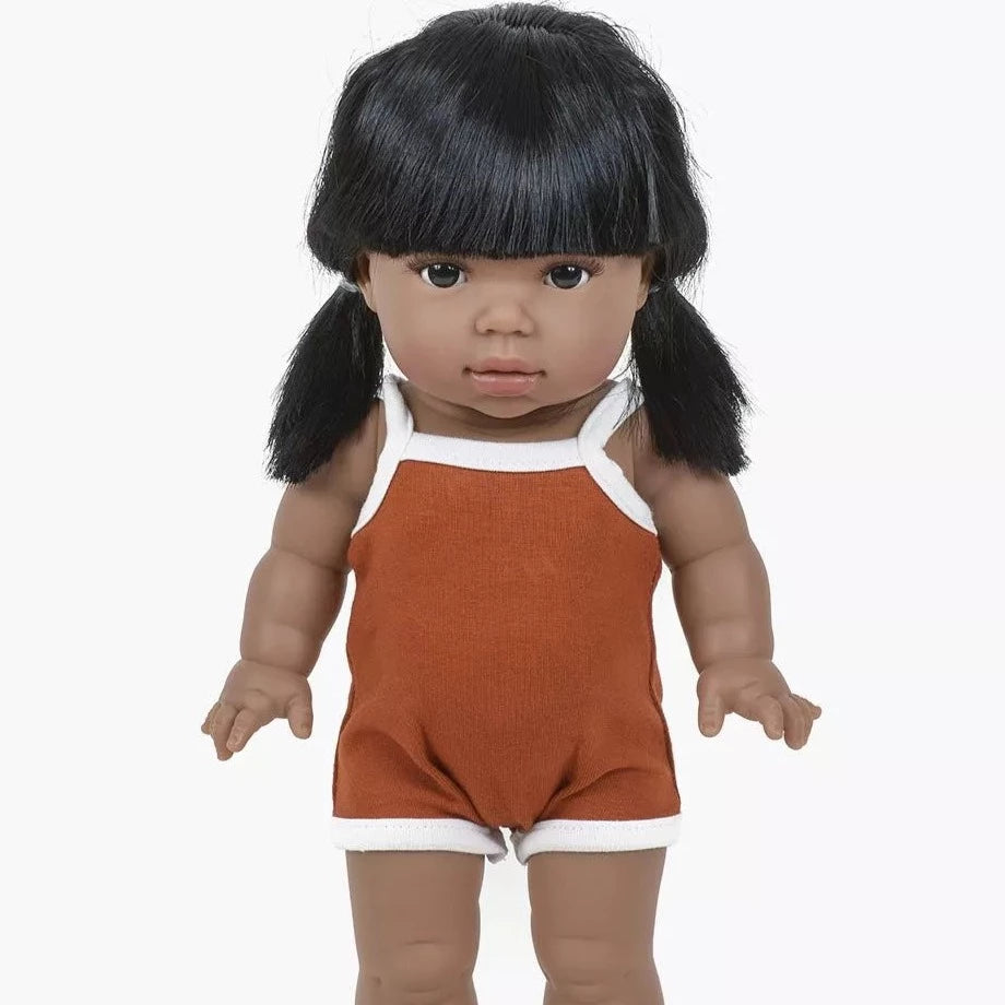 White background with Lika Doll by Minikane. Doll is an indigenous girl with dark black hair, and dark eyes, wearing a spaghetti strap romper in rust.